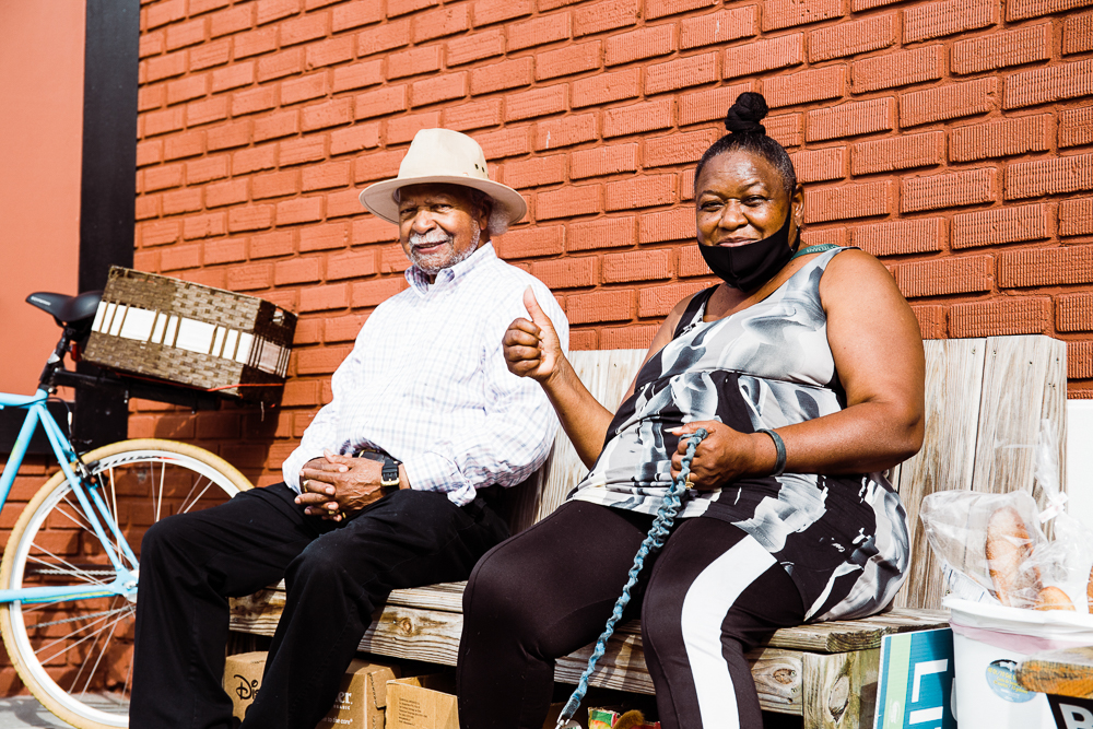 A man and woman sitting on street bench, smiling and giving a thumbs up.
